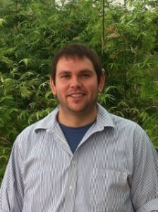 A man with brown hair is smiling and standing in front of a green leafy background. He is wearing a light blue shirt over a dark blue t-shirt.